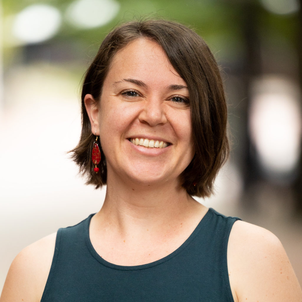 Woman in headshot in green tank top in front of trees, smiling and looking at camera