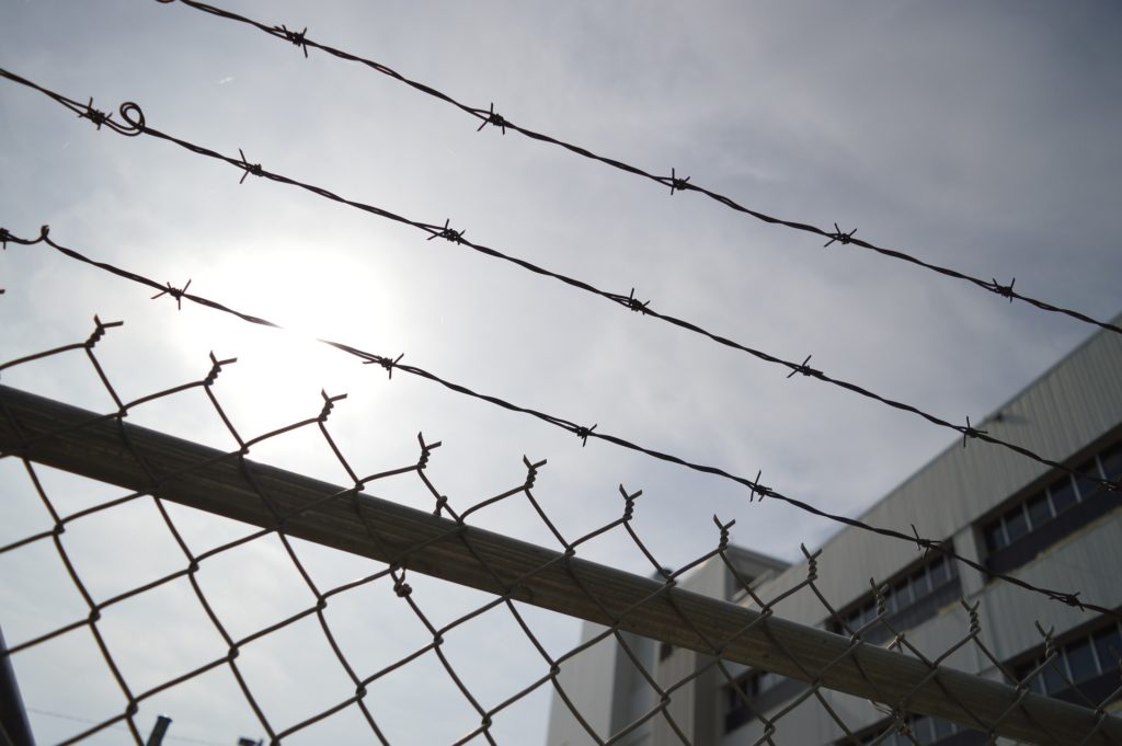 A chain link fence with barbed wire is shown in the foreground with a white building behind it.