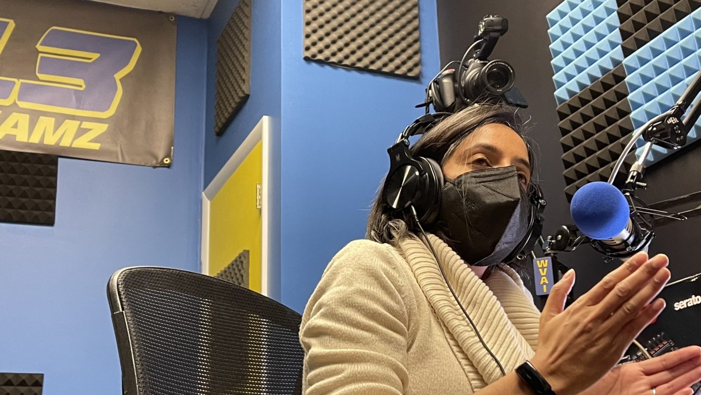 Woman in a beige sweater and black face mask in headphones talks at the microphone in a studio, with a corner of a 101.3 Jamz banner in the background.