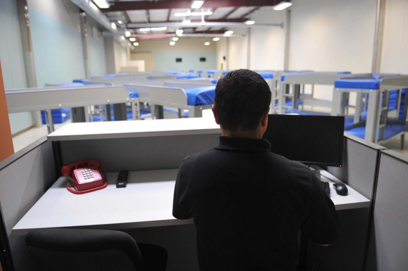 A man stands at a workstation, his back to the camera, with a monitor and red phone on a gray desk. Beyond there are many bunk beds in star patterns with blue mattresses.