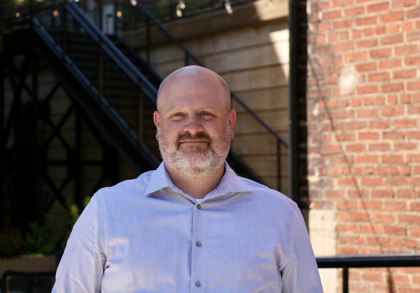 A man in a button downed shirt stands in front of a brick wall.